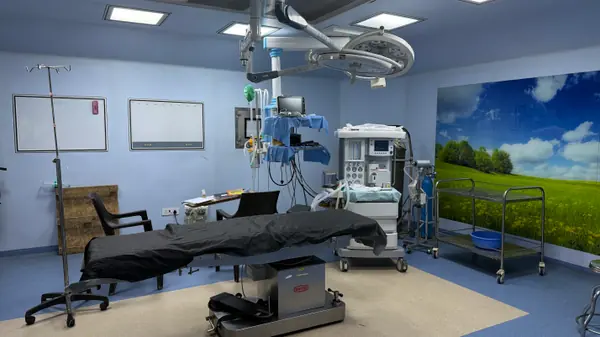An operation theatre with the table under a twin-dome surgical light, an anaesthesia machine and a monitor beside it.