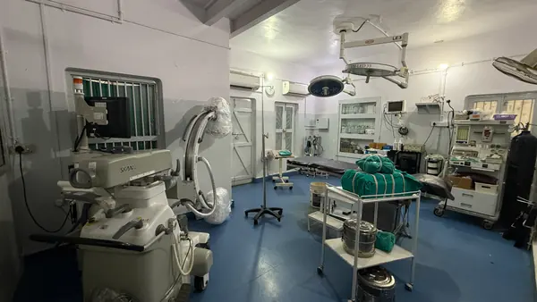 A procedure room with a mobile C-arm, an overhead surgical light, an anaesthesia trolley and wrapped sterile instrument sets.